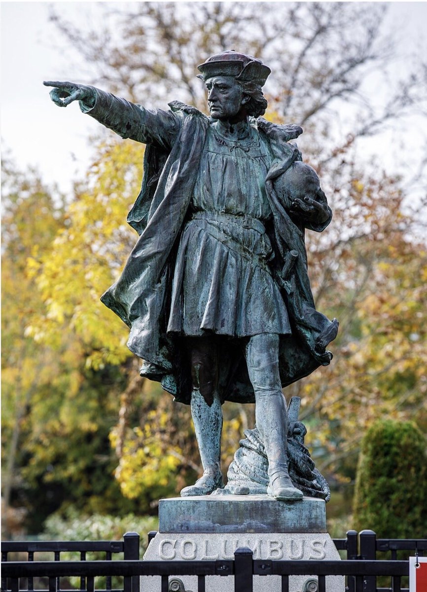A bronze statue of a historical-looking man wearing a cloak and hat stands outdoors, pointing to the left and holding a helmet. Trees with autumn foliage are in the background, and the statue stands on a stone pedestal with text.