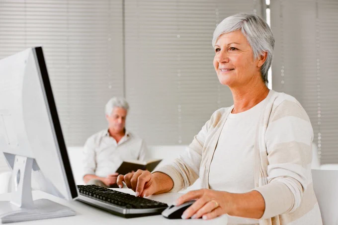 Mature woman smiling and focused while using a desktop computer in a bright office setting, with a man reading in the blurred background.