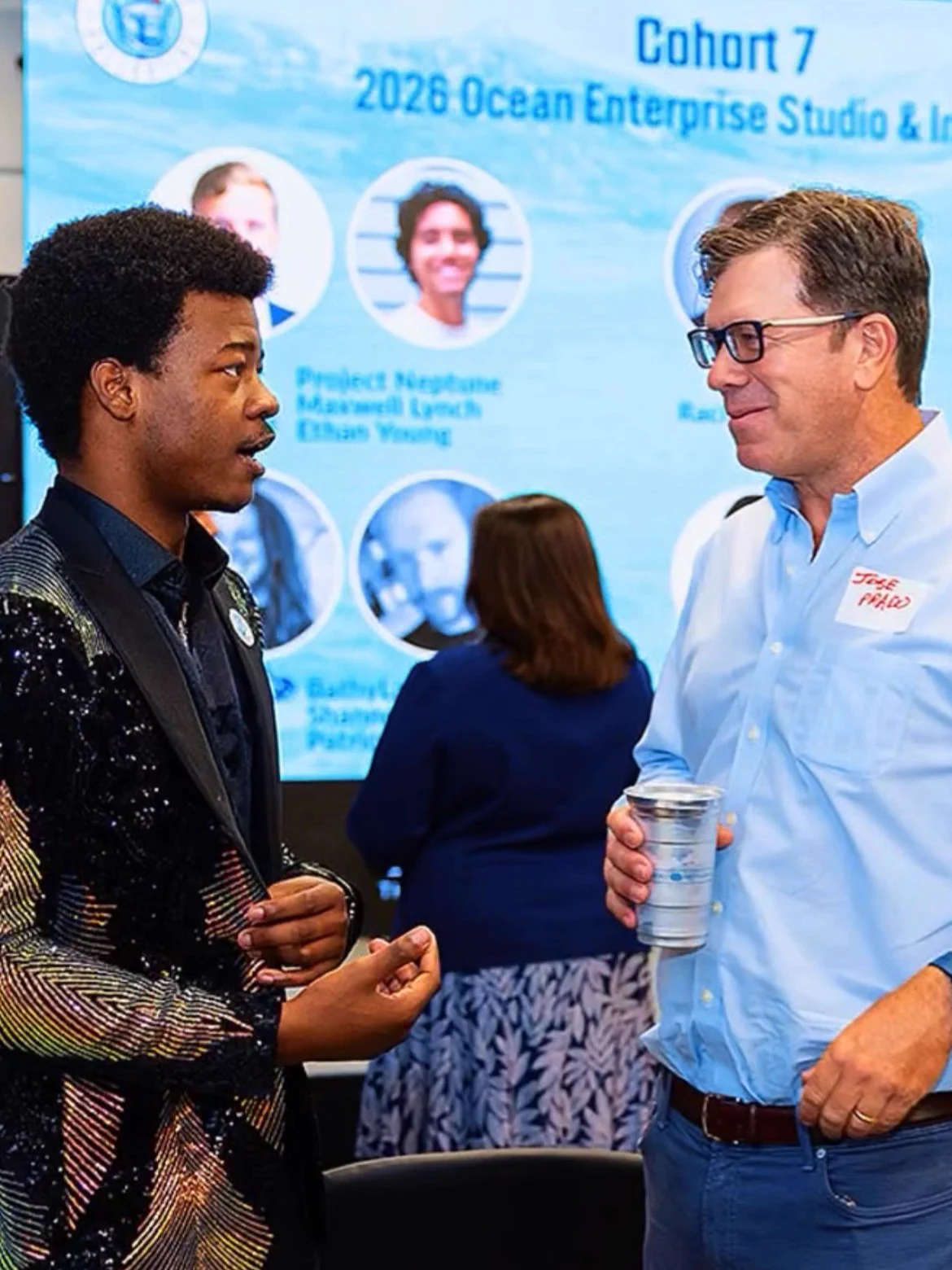 Two attendees speak at Seaworthy Collective’s startup showcase. The person on the left gestures while the man on the right listens, holding a cup. A screen behind them displays the Ocean Enterprise Studio & Incubator’s Cohort 7.