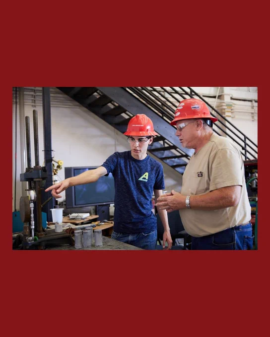 Two people in safety gear, including red hard hats and goggles, stand in an industrial setting. One person points at equipment on a table, while the other observes. Stairs and industrial machinery are visible in the background.