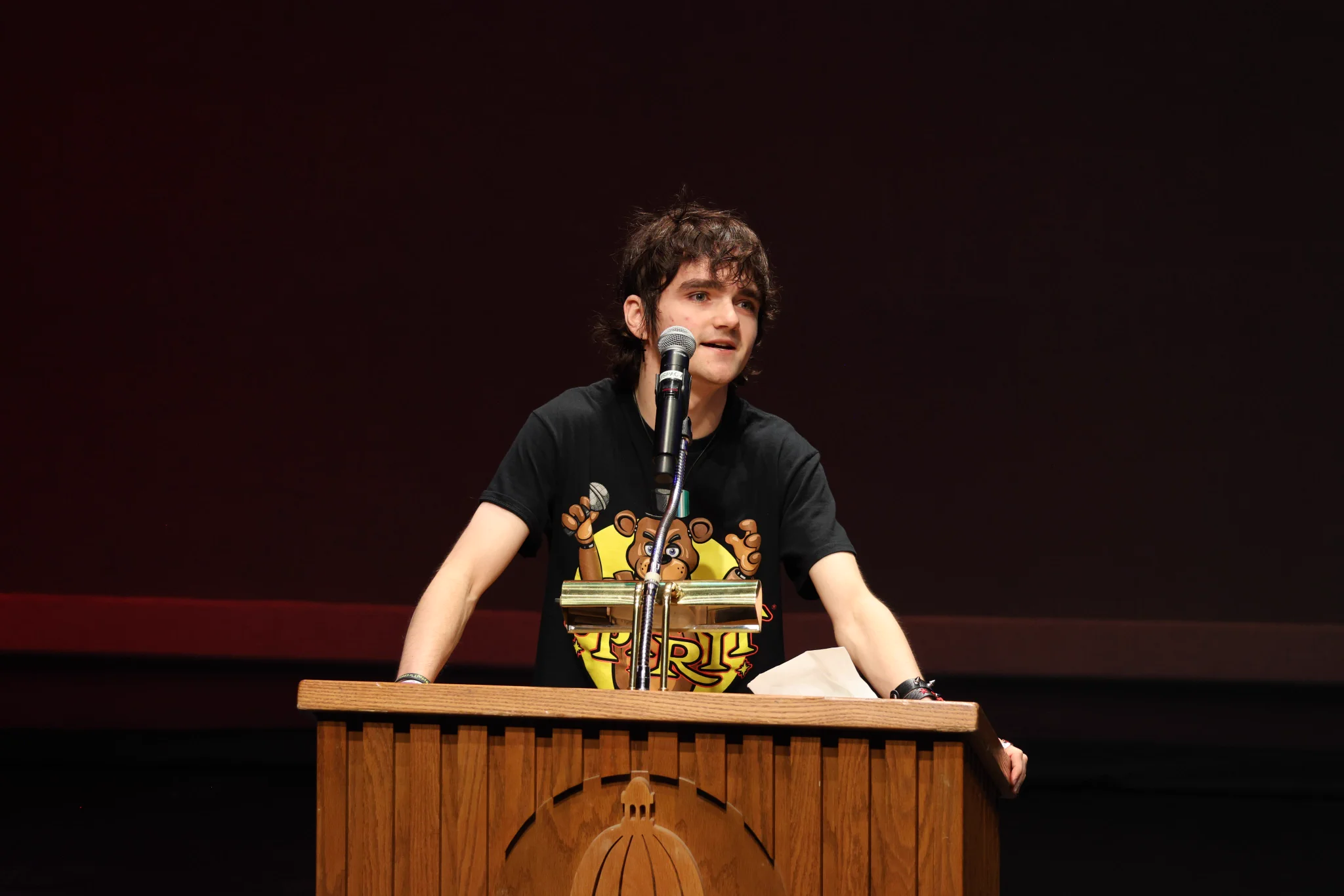 A young man in a graphic T-shirt leans on a wooden podium and speaks into a microphone.