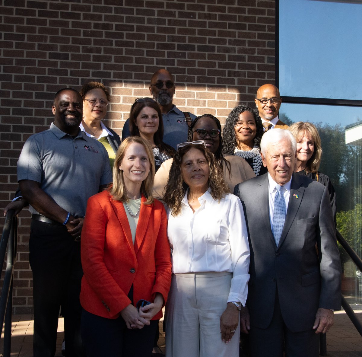 A group of adults pose for a photograph outside a brick building, standing beside a large window. There is no prominent readable text.