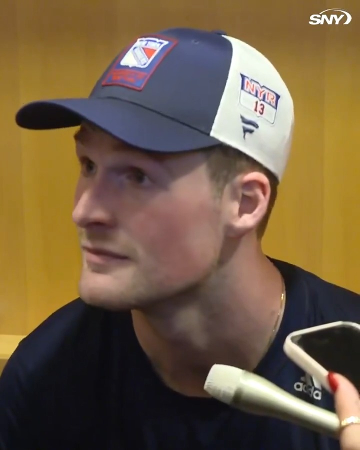 Alexis Lafrenière, wearing a New York Rangers cap marked "NYR 13" and a fan's #13 jersey, speaks to reporters during a postgame interview following his 7-goal performance over a 5-game span, smiling as he reflects on the team's strong play and enthusiastic home crowd.