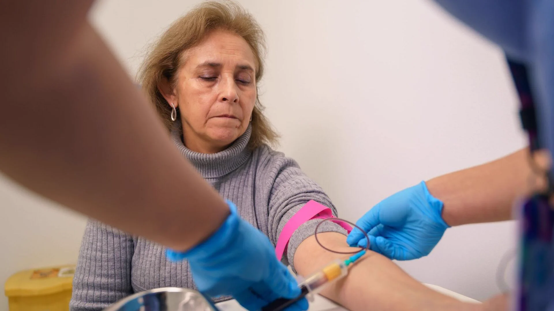 A person with closed eyes is having blood drawn from their arm by a healthcare professional wearing blue gloves.