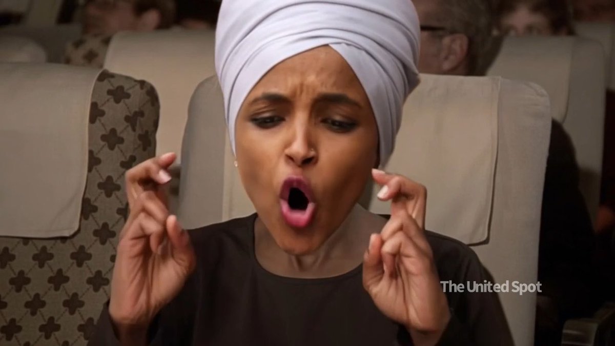 A woman wearing a light-colored turban reacts with an exaggerated surprised expression, mouth open and fingers crossed, on an airplane; watermarked "The United Spot".