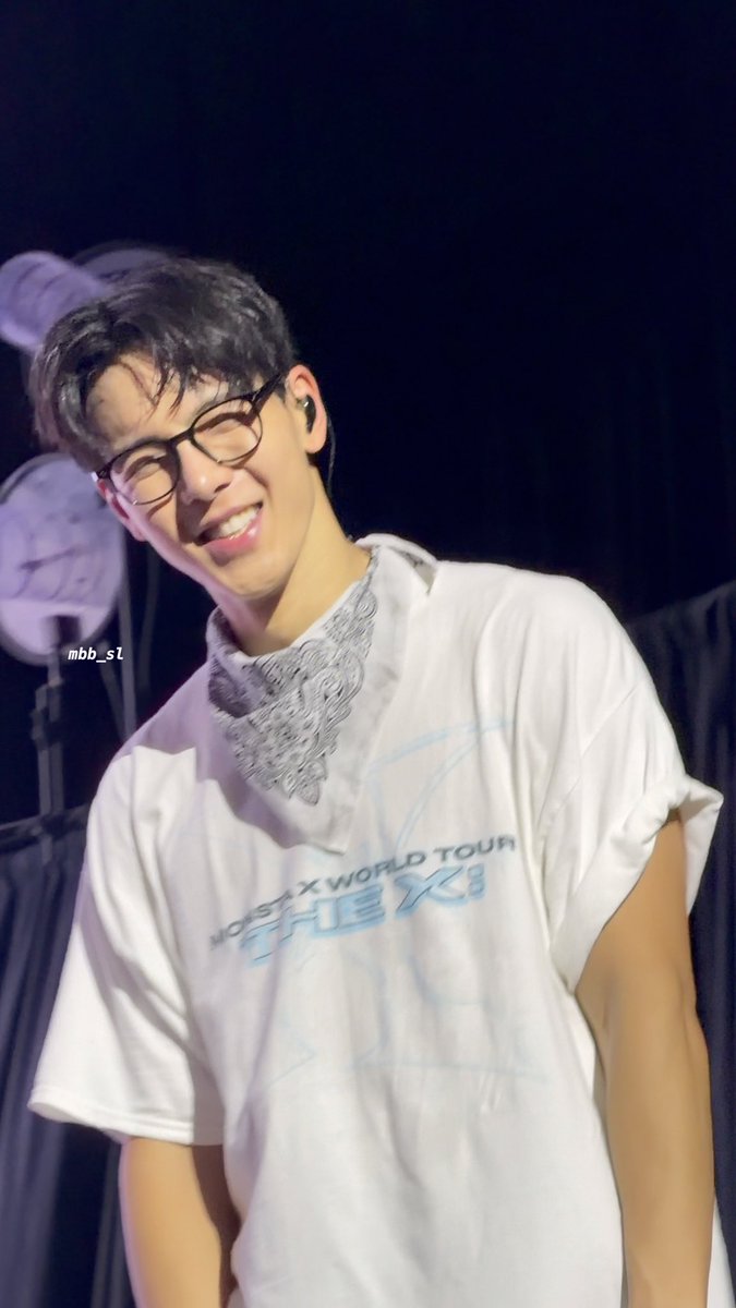 A young male performer with glasses smiles on stage, wearing a white "MONSTA X WORLD TOUR" t-shirt and a black patterned bandana around his neck.