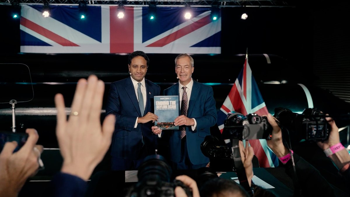 Two men in suits stand together on a stage holding a sign reading "REFORM UK FINDING THE ASYLUM SCAM," with a British flag behind them and photographers in the foreground.