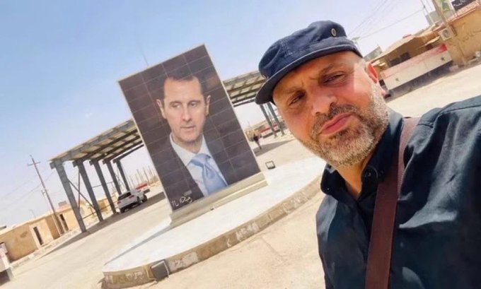 A man wearing a dark cap and shirt takes a selfie in front of a large outdoor portrait of Syrian President Bashar al-Assad in a public square. The image relates to the post about Syrian intelligence operations by visually anchoring the subject to the Assad regime, the authority conducting the arrest of terrorist militia leader Ahmed Hassan Al-Khafaaji (Abu Tabarak).