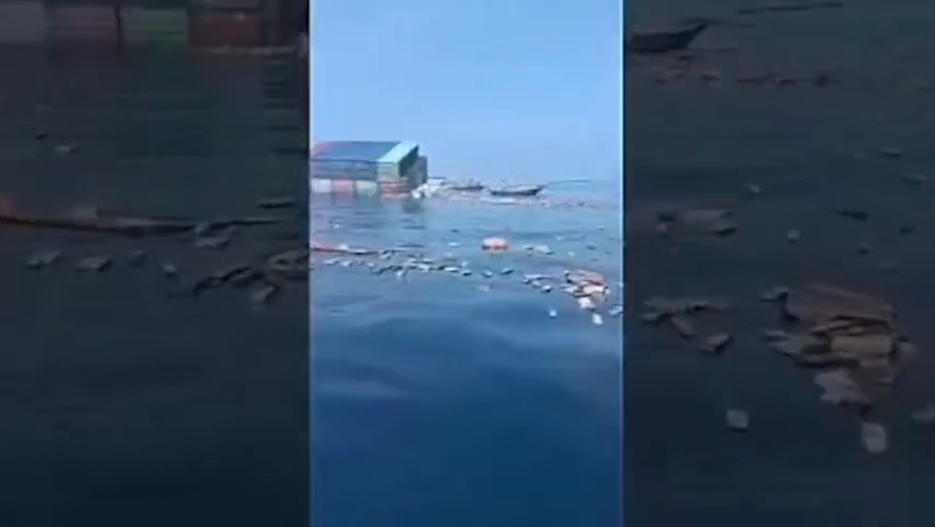 Vertical video footage of floating debris and objects scattered across a body of water near what appears to be a structure or shoreline.