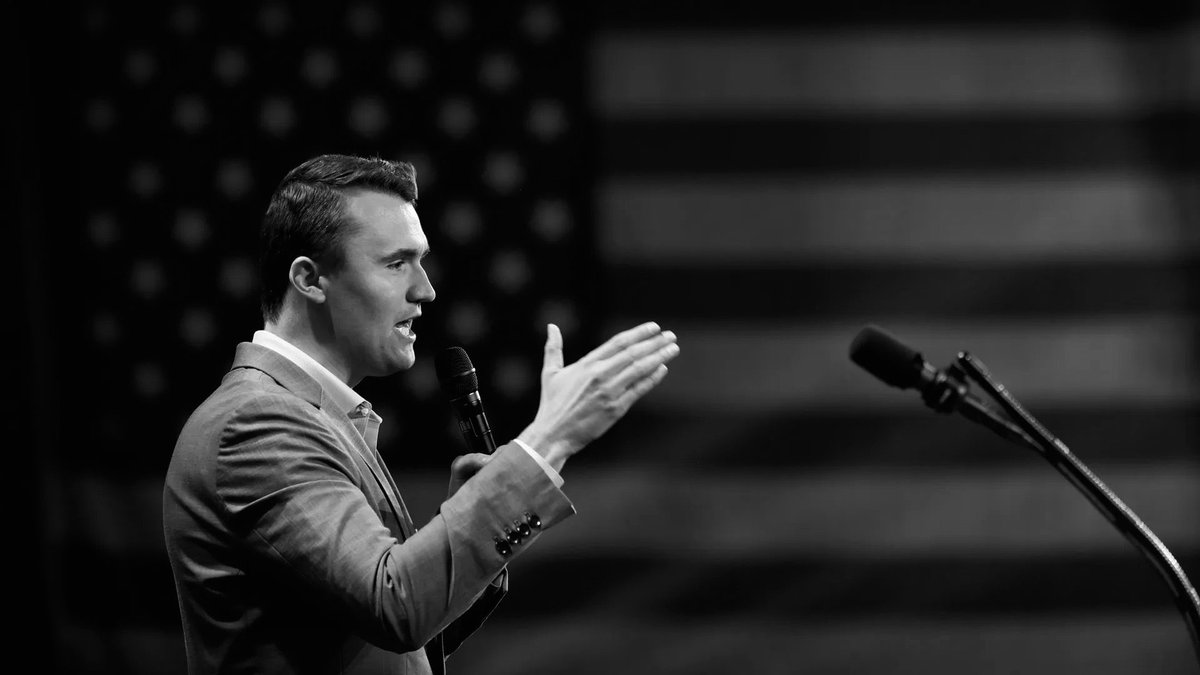 A black-and-white photograph of a man in a suit speaking into a handheld microphone and gesturing onstage. A second microphone on a stand is visible to the right, with a blurred background. There is no clearly readable text.