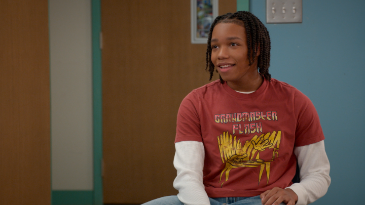 A young man with braided hair smiles warmly while wearing a red "Grandmaster Flash" graphic t-shirt layered over a white long-sleeve shirt, seated in what appears to be an office or classroom setting with a light switch panel visible on the wall behind him. His friendly, approachable expression matches the cute sentiment expressed in the post.