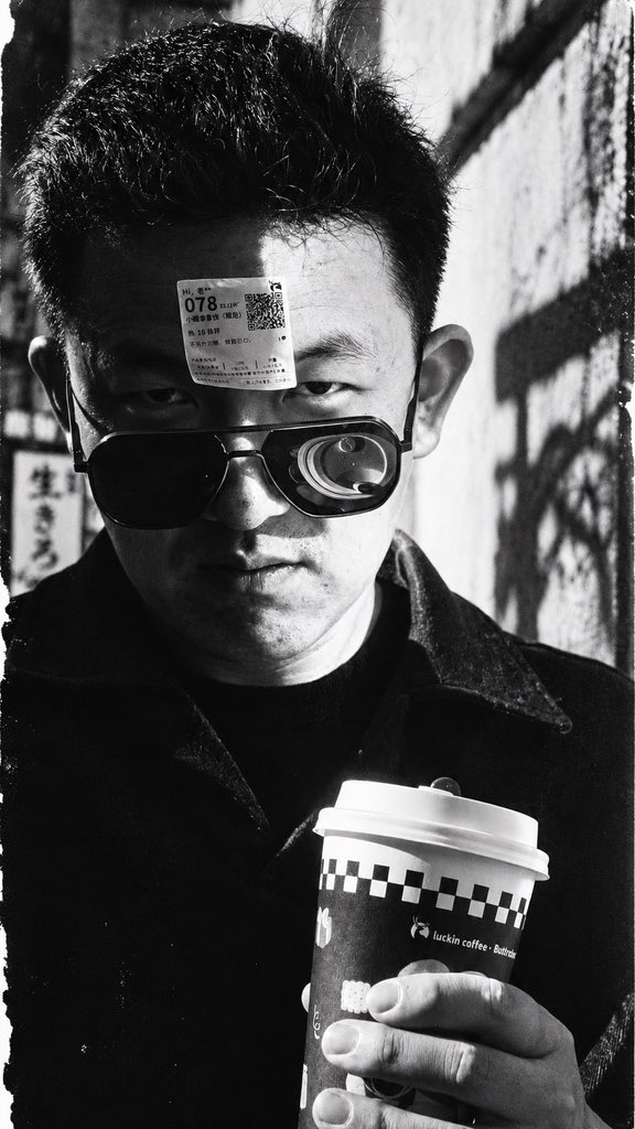 zeallaez_'s tweet photo. 当AI可以创造一切，我们为什么还要亲自创造？

今天，一条很久以前的帖子突然收到了回复。

原帖大概是在感叹，AI出现以后，摄影可能要消失了。博主展示了许多AI生成的美女照片，那些曾经需要摄影师、模特、灯光、场景和后期才能完成的糖水片，如今只需要输入几句话，就能轻易获得。

我当时在评论区留下过一个观点：这反而凸显了摄影师的重要性。

当然，这里说的重要性，未必是商业上的。倘若摄影的全部价值只是一张漂亮的照片，那么AI确实已经让许多传统摄影工作失去了存在的理由。

但我始终觉得，一张照片和拍摄一张照片，是两回事。

你走进一条陌生的街道，等待光线变化，与一个陌生人交谈，观察他的神情，甚至因为某个偶然出现的瞬间，改变了原本的拍摄计划。那些气味、声音、温度、情绪，以及你在现场接收到的复杂信息，最终可能只有极少一部分留在照片里。

然而，真正改变你的，恰恰可能是那些没有被记录下来的东西。

今天重新看到这条评论，突然又想起最近读到的一些关于人机共创的讨论。

其中一个中国学者与AI共同探索经济学新概念的案例，让我对所谓的“共创”有了更具体的理解。

一个经济学家，无论多么博学，都无法摆脱自身经验、知识结构和时代环境的局限。AI同样有它的局限，它可以组织庞大的信息，建立看似遥远的关联，却未必真正理解一个人为什么会对某个问题感到困惑。

有趣的事情发生在两者相遇之后。

人提出一个模糊的直觉，AI给出一种意想不到的解释；人发现其中的漏洞，又重新组织自己的问题。如此反复，一些原本不可能独自抵达的想法，或许就这样慢慢浮现出来。

当然，这种碰撞未必产生真正的新知识，也可能只是彼此强化偏见。但它至少提供了一种可能：创造不再只是一个孤独的头脑向外输出思想，也可以成为一个不断受到外部刺激、质疑和改变的过程。

我忽然意识到，这与摄影竟然有某种相似之处。

摄影师与现实相遇，思想者与AI相遇。真正有价值的，可能都不是最终得到的那个结果，而是相遇过程中发生的变化。

于是，一个更令人不安的问题出现了。

如果有一天，AI不仅比我们拍得好、写得好、设计得好，甚至比我们更擅长提出问题，那么人为什么还需要亲自创作？

过去，我们似乎总是习惯用结果衡量创造。一本书、一张照片、一栋建筑、一套理论，仿佛只有留下某种可以展示、交换和评价的成果，创造才算完成。

但一个人为什么要弹钢琴？如果只是为了听到美妙的音乐，打开播放器显然更有效率。一个人为什么要画画？如果只是为了获得一幅漂亮的图像，机器可能早已做得更好。

我们仍然愿意去做这些事情，也许只是因为，在那些笨拙、缓慢、充满不确定性的过程中，我们感受到自己正在活着。

就像今天这张照片。

原本只是阳光下随手拍的一张自拍，额头贴着咖啡标签，戴着墨镜，拿着一杯咖啡，带着一点无聊的戏谑。我让AI把它处理成粗粝的黑白纪实影像，阴阳脸、强烈的反差、浓重的颗粒。

同一个人，同一个瞬间，忽然呈现出一种完全不同的情绪。

我开始好奇，这种情绪究竟原本就藏在照片里，还是AI替我发现了另一种理解自己的方式？

也许这才是我真正感兴趣的AI创作。

我并不需要它替我生产无数张漂亮的照片。我更希望它能让我重新观看那些习以为常的事物，甚至让我对自己原本确信的判断产生怀疑。

技术越强大，这种愿望反而越迫切。

因为我们完全可能进入这样一个时代：一个人拥有前所未有的表达能力，却越来越少拥有真正想表达的东西；可以轻易获得无数答案，却逐渐失去提出问题的欲望。

倘若连自己的好奇、热爱、痛苦和欲望，都习惯性交给机器安排，那么即使我们拥有了整个世界的知识，又究竟剩下多少属于自己的生活？

人机共创的意义，或许正在这里。

它不必证明人类永远比机器特殊，也不必执着于捍卫某种不可替代的能力。它只是提醒我们，技术可以替代许多劳动，却没有理由要求我们放弃亲自经历世界的愿望。

人活着究竟有什么意义？这个问题困扰了哲学家几千年，至今也没有一个令人满意的答案。

但我越来越觉得，意义也许从来不是某个等待我们找到的最终答案。

它可能就发生在一次偶然的拍摄、一场没有预料到的谈话、一个突然冒出来的念头里。

我们创造世界，也在这个过程中不断创造自己。

而这件事，我暂时还不想完全交给AI。