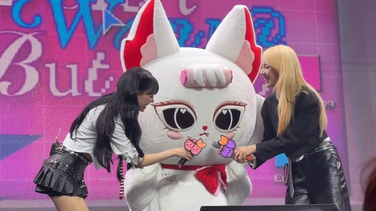 Two women interact with a large white cat-like mascot costume on a stage with a pink backdrop, with one holding a "My Little BUT" sticker.