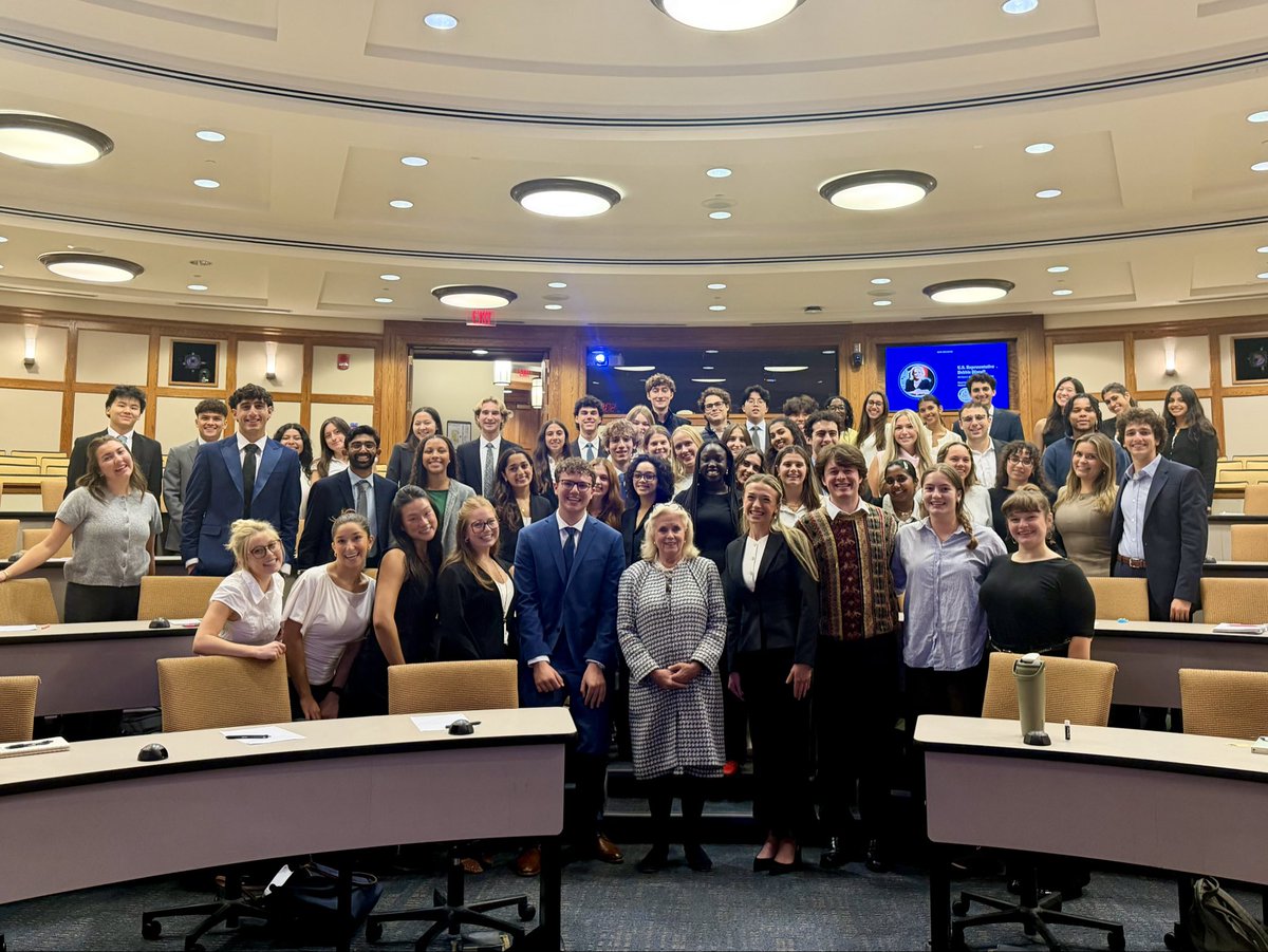 A large group of people, mostly young adults, pose together in a classroom or lecture hall. Many are dressed in formal or business-casual clothing. Text is visible on a wall display and above a doorway.