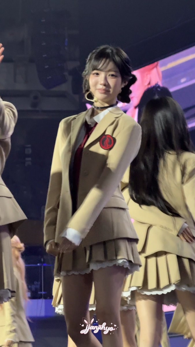 Performance photo of a young female K-pop idol wearing a tan school uniform blazer, red tie, and pleated skirt on stage, with other performers visible in the background.