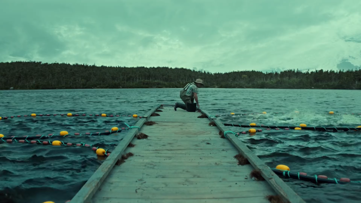 A person wearing a hat kneels on a wooden dock extending over a greenish lake, with floating aquaculture lines containing yellow and red buoys.