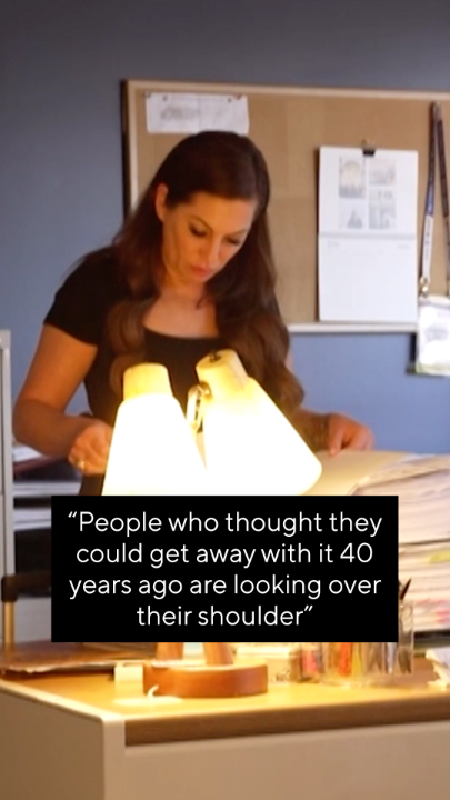A woman reviewing documents at a desk with a lamp, with text overlay reading: "People who thought they could get away with it 40 years ago are looking over their shoulder".