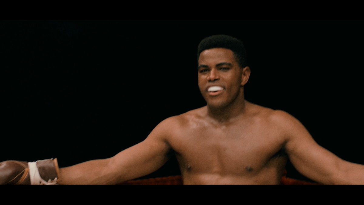 A dramatic still of a shirtless Muhammad Ali wearing boxing gloves in a boxing ring against a dark background.