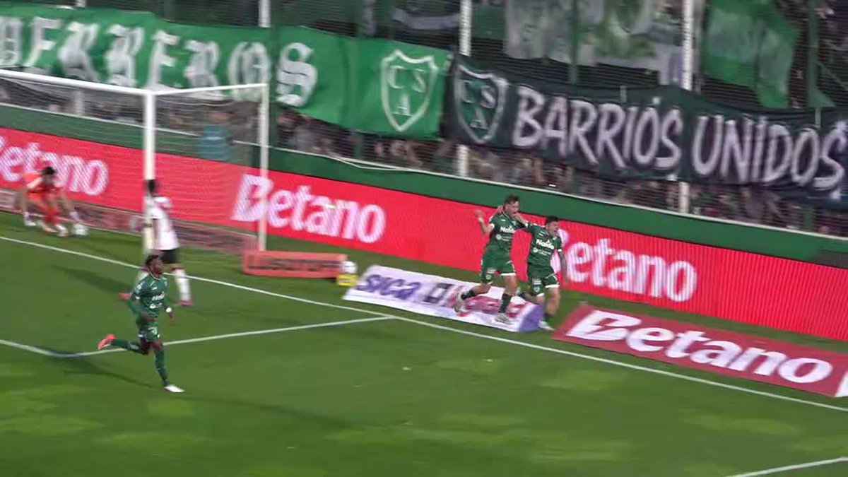 Soccer players in green uniforms celebrate a goal near the goalpost during a match, with 'BARRIOS UNIDOS' banners and 'Betano' advertising boards visible in the background.