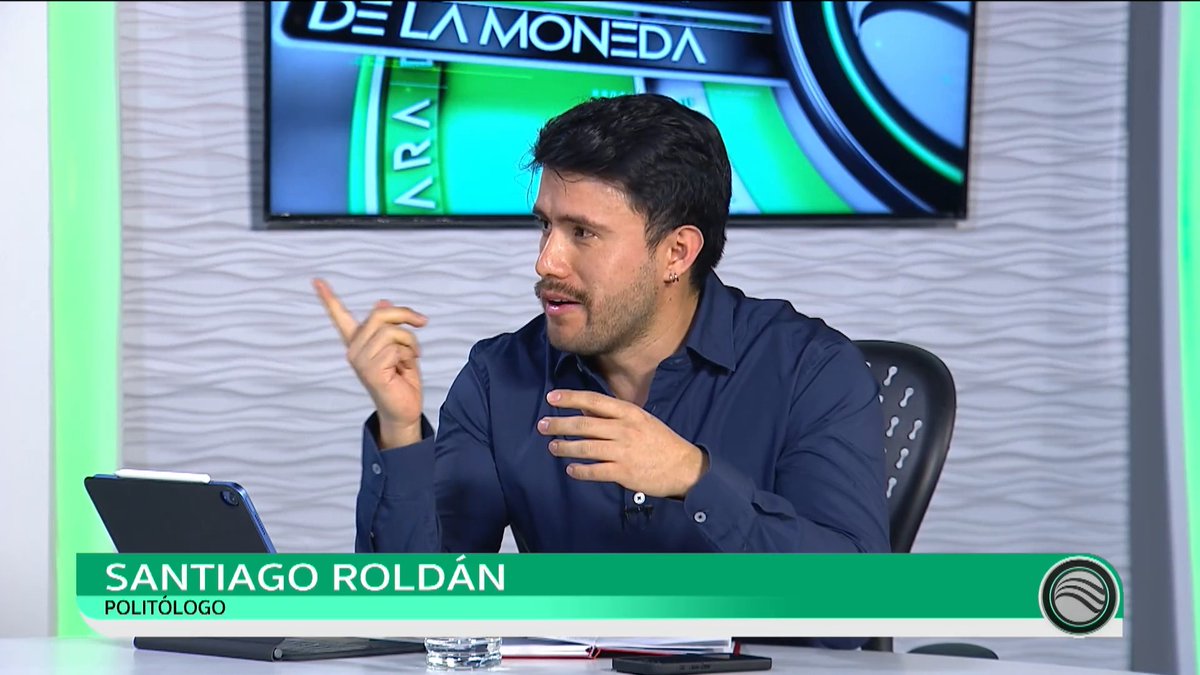 A television broadcast still showing politologist 'Santiago Roldán' speaking and gesturing on a show set, with 'DE LA MONEDA' visible on a screen behind him.