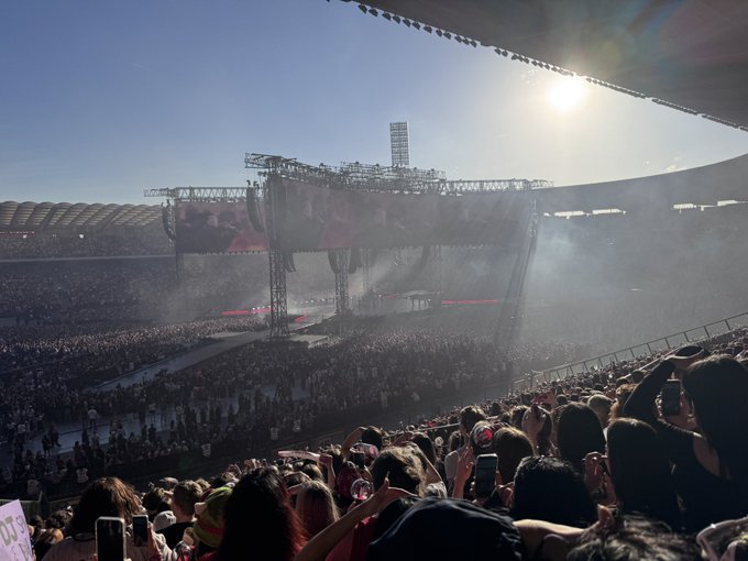 A large concert stadium is filled with a massive crowd under bright sunlight, with fans holding up illuminated fan signs and a large video screen above the stage.