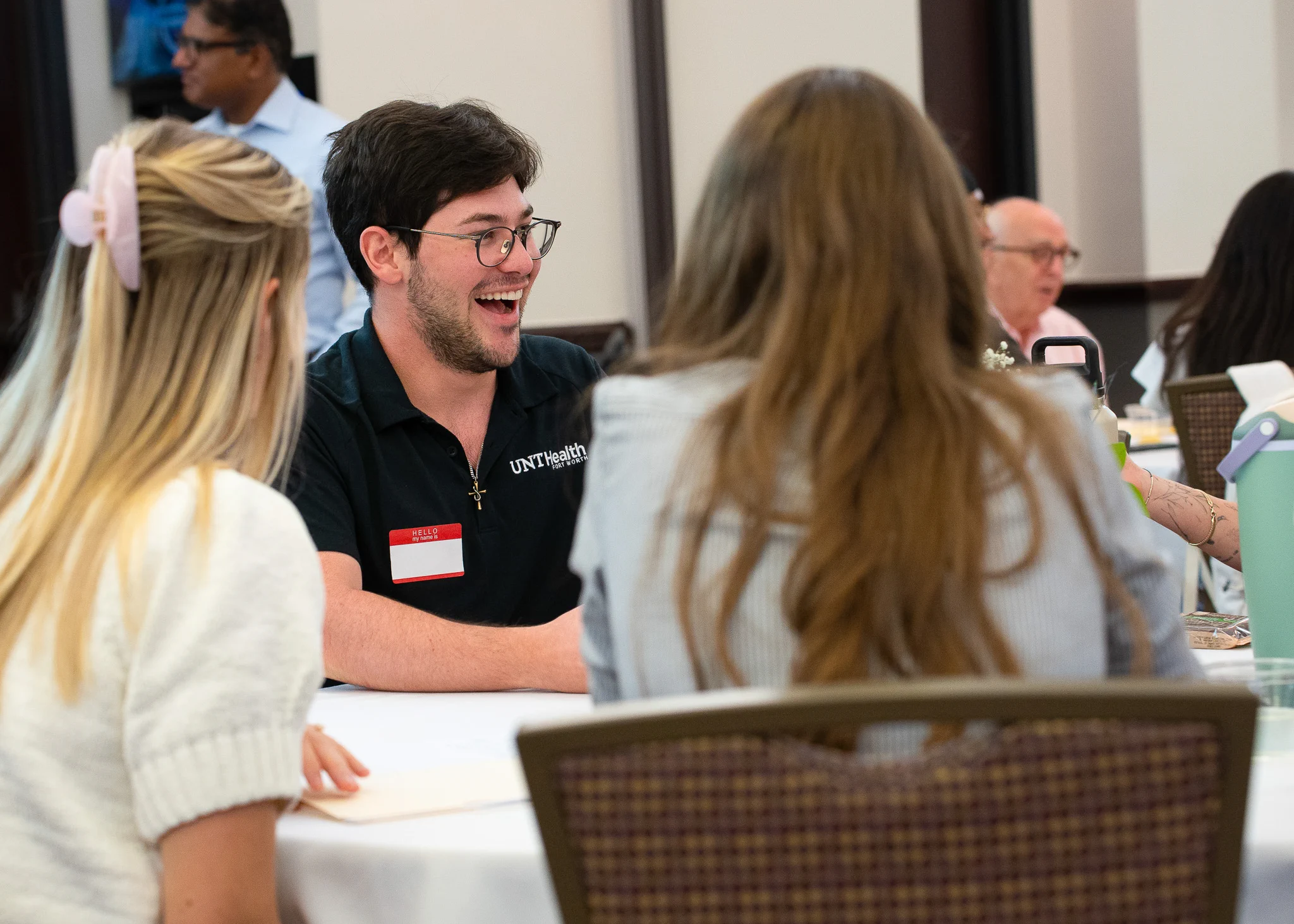 A group of people sitting at a table in a conference setting, engaged in conversation. The person facing the camera is wearing a shirt with a logo that says "UNT Health." Others around the table are listening and interacting. There are glasses and papers on the table.