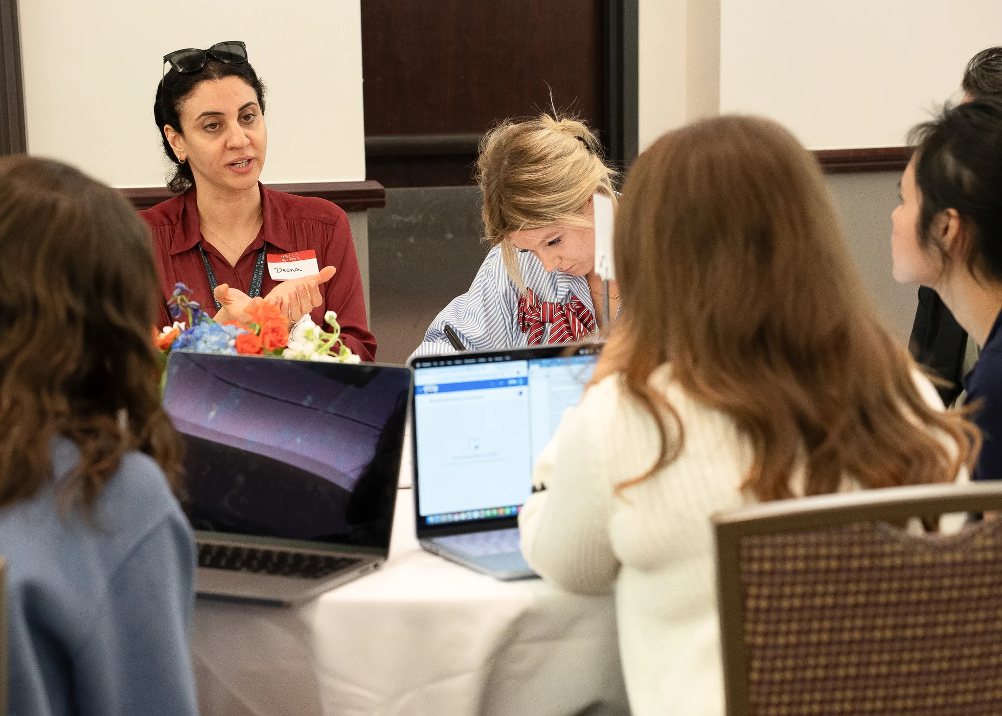 A group of individuals seated around a table, engaged in discussion. Laptops are open in front of some participants. One person wearing a red shirt is speaking, while others listen attentively.