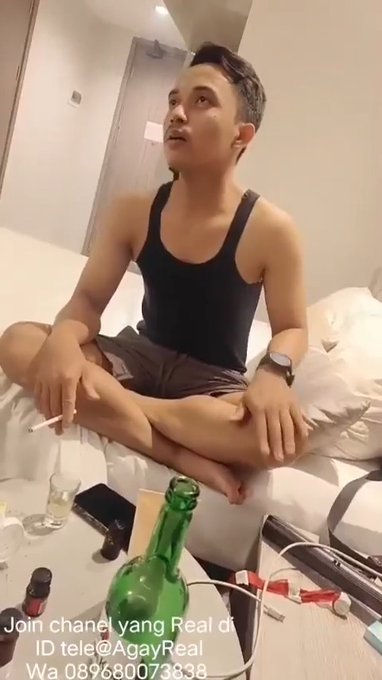 A young man in a black tank top and shorts sits cross-legged on a bed, holding a cigarette, with a green bottle and various items scattered on the table in front of him. Text overlay promotes a Telegram channel "@AgayReal" and contact number "089680073838," suggesting the post is advertising social media or messaging services, with the caption "mas singlet hitam" (Indonesian for "black tank top guy") referencing his outfit.