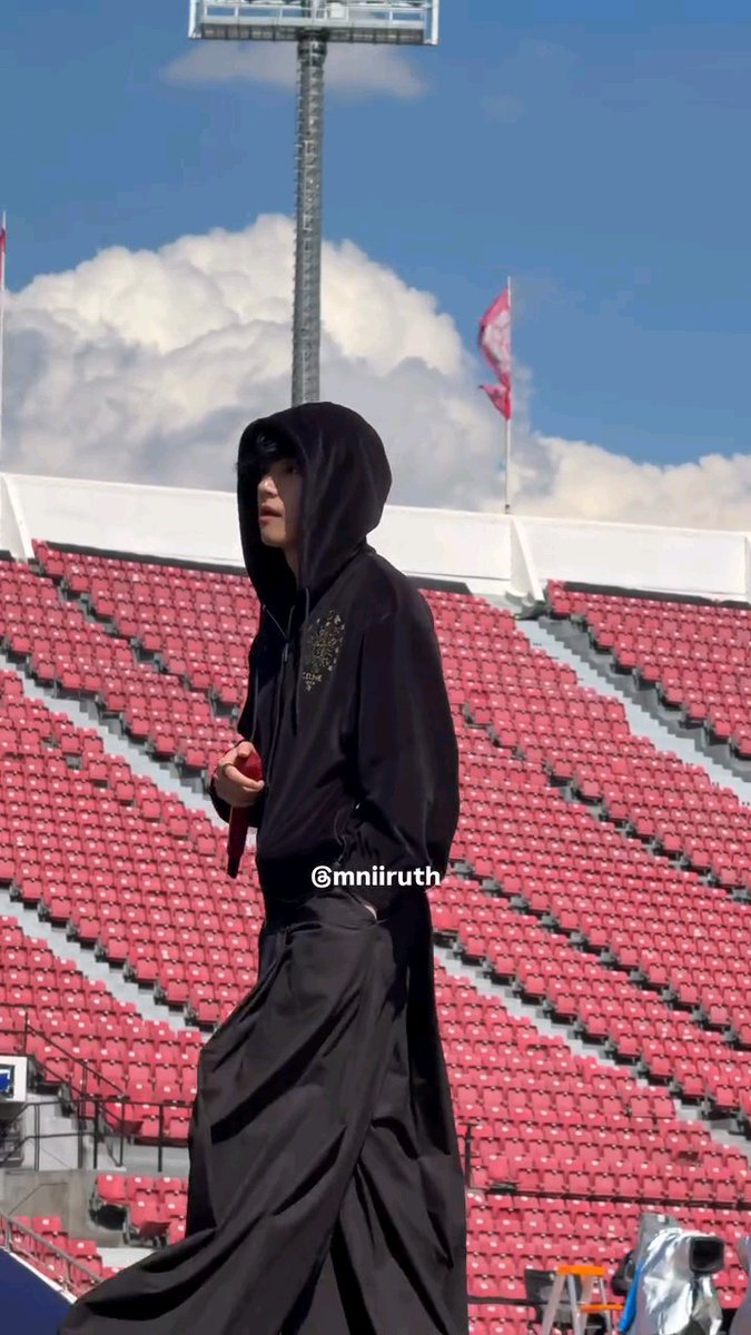 A person wearing a long black hooded robe stands in an empty stadium with red seats, with the watermark "@mniiruth" visible.