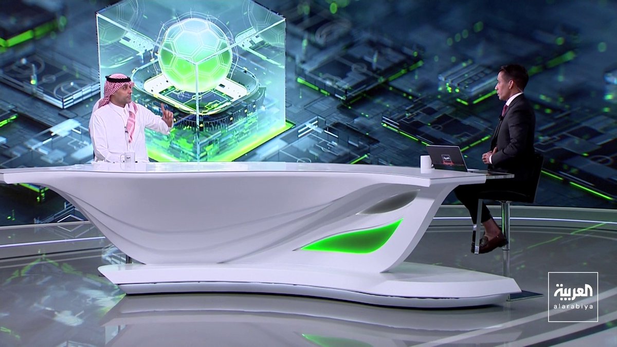A television news studio set on Al Arabiya channel features a modern white desk with green accent lighting, where two men sit facing each other—one dressed in traditional Saudi attire (white thobe and red-checkered keffiyeh) gesturing while speaking, and another in a dark suit with a laptop. Behind them, a large digital backdrop displays a glowing green holographic soccer ball inside a transparent cube, reflecting the sports broadcast theme of the program "Fil Marma" discussing Saudi football. The Al Arabiya logo appears in the bottom right corner of the screen.