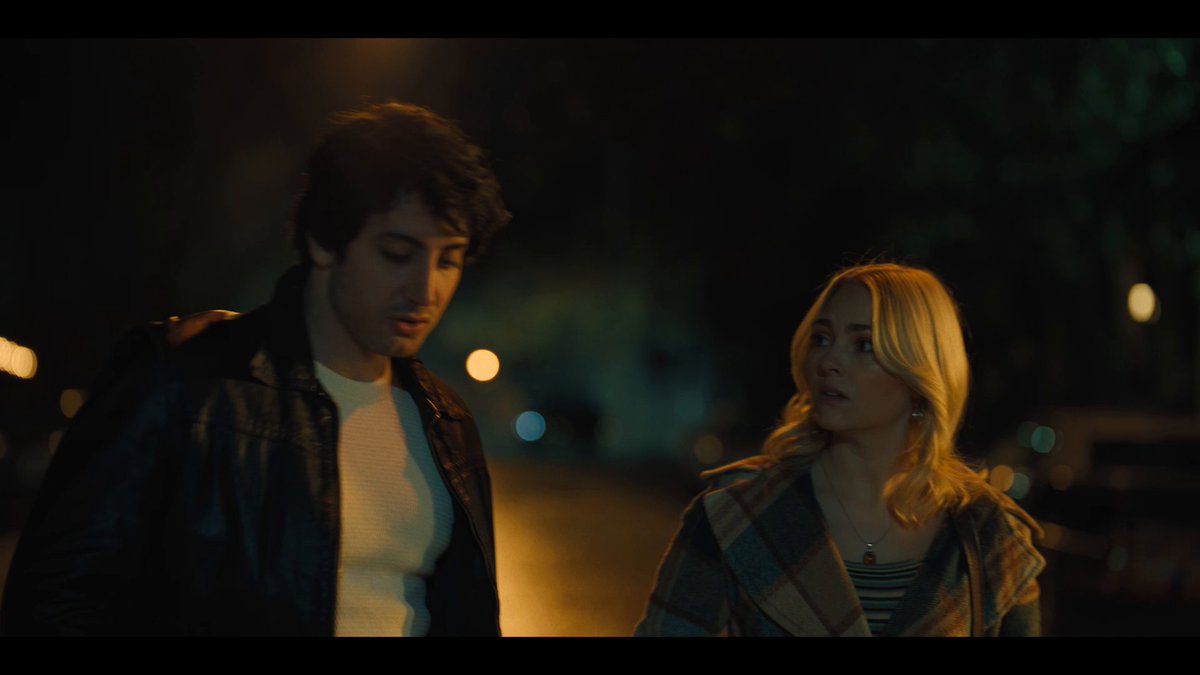 A nighttime scene from what appears to be a film, showing a young man in a leather jacket and a blonde woman in a plaid coat walking together on a dimly lit street.