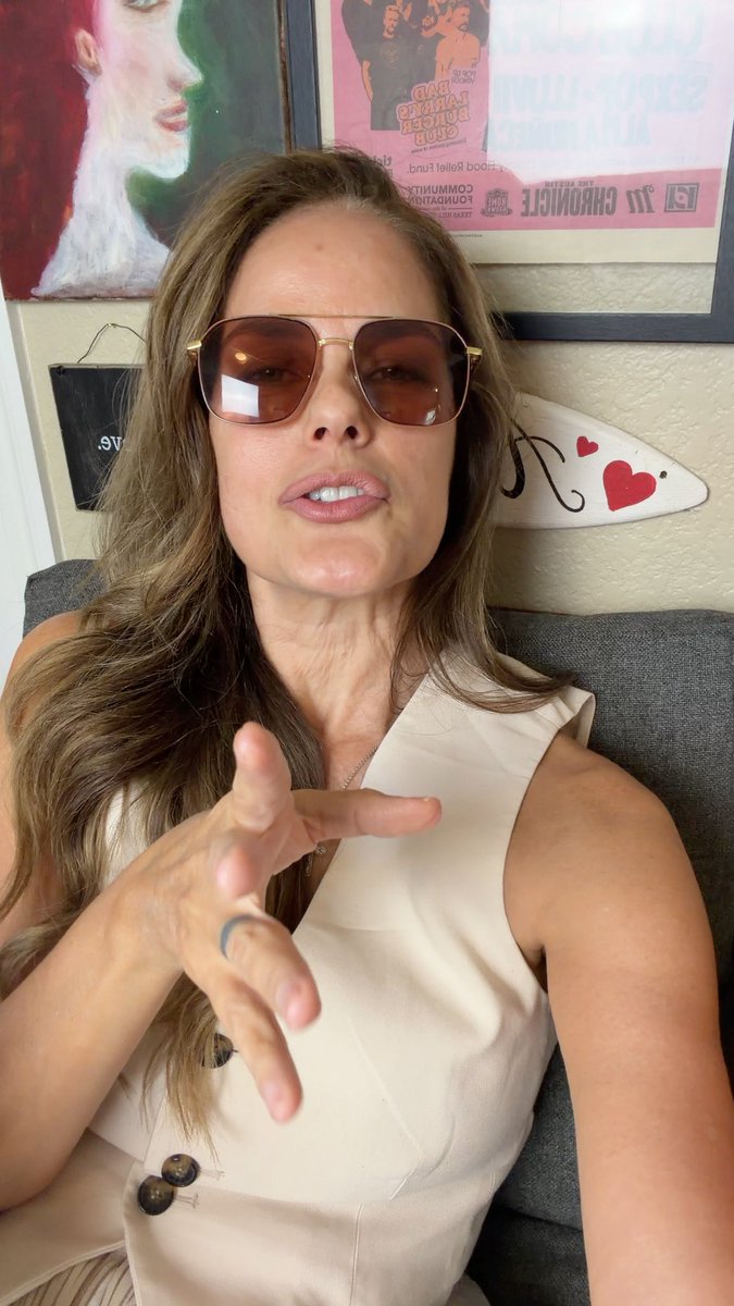 A woman wearing brown-tinted aviator sunglasses and a beige sleeveless top gestures with her hand, sitting in front of framed artwork and a decorative surfboard.