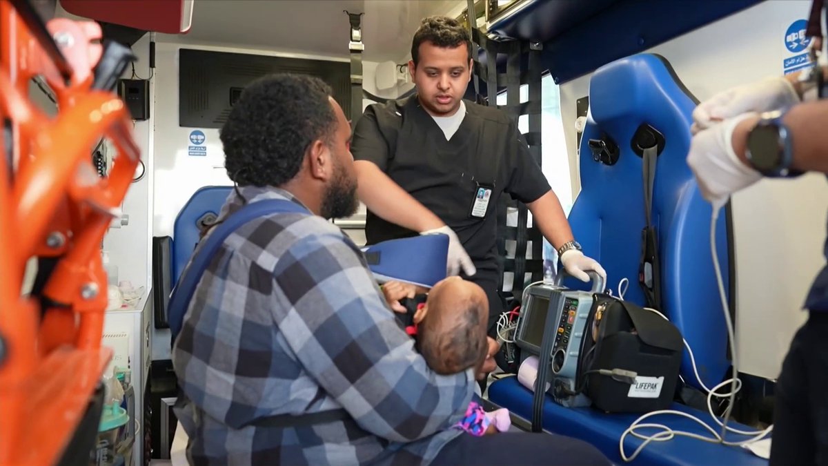 A paramedic in a dark uniform attends to a patient seated inside an ambulance while another person holds a baby, with medical equipment visible including a LIFEPAK monitor.