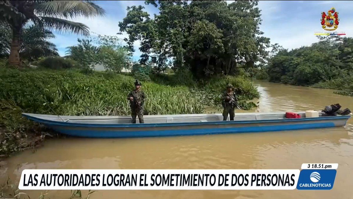 Two soldiers in uniform stand armed in a blue and white wooden boat on a muddy river, surrounded by lush tropical vegetation. The news broadcast from Cable Noticias features Spanish text reading 'LAS AUTORIDADES LOGRAN EL SOMETIMIENTO DE DOS PERSONAS' with a timestamp of 3:18:51 pm.