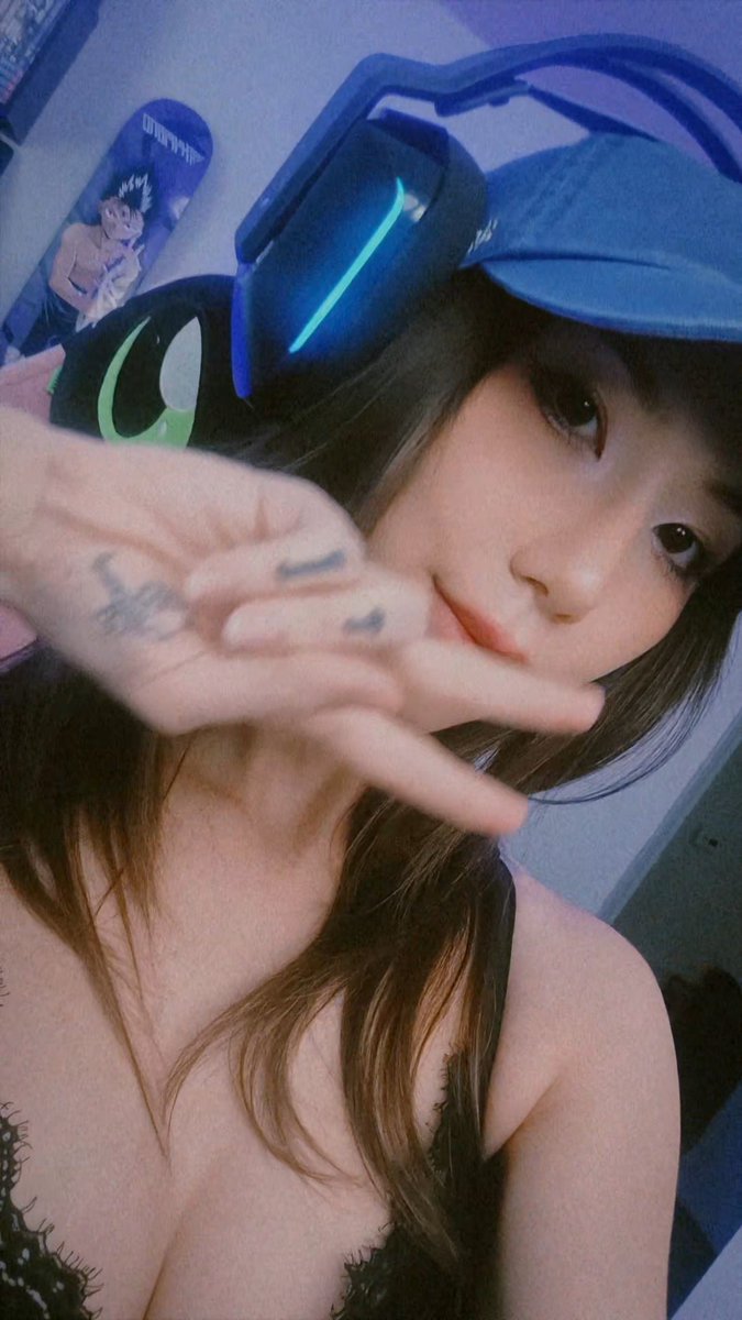 A young woman wearing blue cat-ear headphones and black lace lingerie poses for a selfie, making a peace sign with her ink-marked hand.