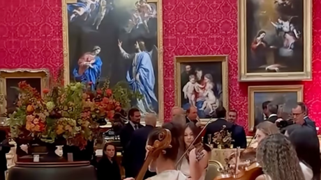 A formal gathering with people seated at a long table, set against a wall covered in red damask wallpaper and adorned with classical framed paintings.