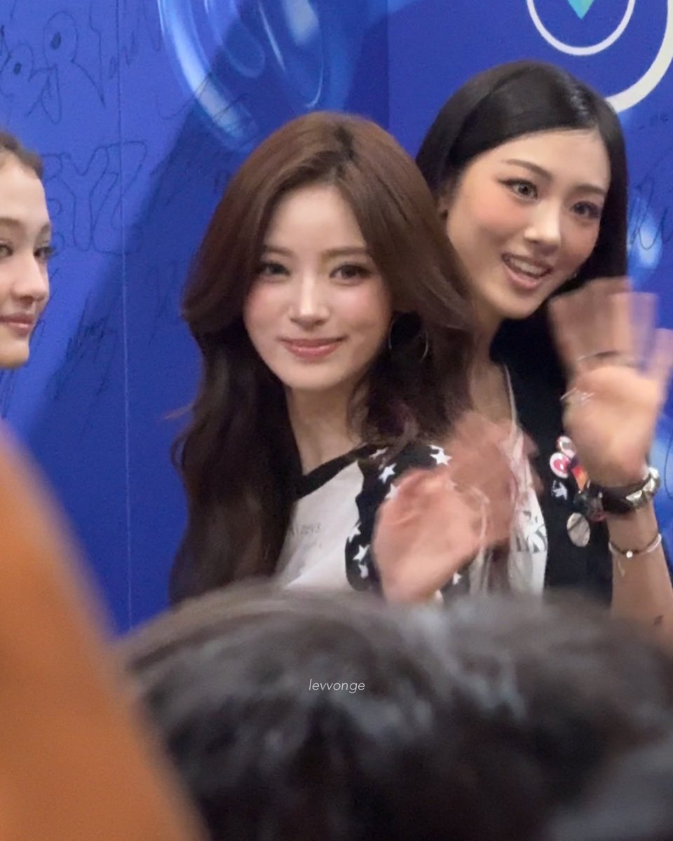 Two women smiling and clapping at what appears to be a public event with a blue backdrop; the woman on the left has long brown hair and wears a star-patterned top, while the image is watermarked "levvonge".