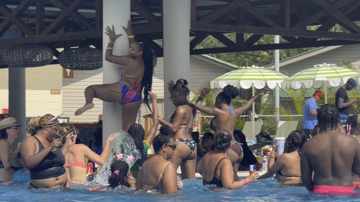 A lively outdoor pool party scene shows many people in swimwear enjoying themselves under a covered pavilion with green and white striped umbrellas in the background.