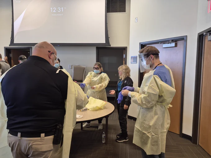 A group of people stand around a table in a room. They are wearing protective gear such as gowns and face shields. A person is talking to them, possibly demonstrating a procedure. A screen is on the wall in the background displaying a digital clock reading "12:31.