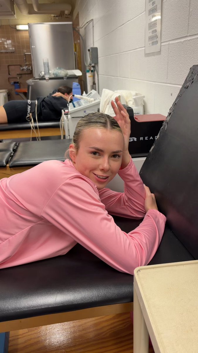 A young woman in a pink long-sleeve athletic shirt lies on a black treatment table in a training room, smiling at the camera while another person lies on a table in the background.
