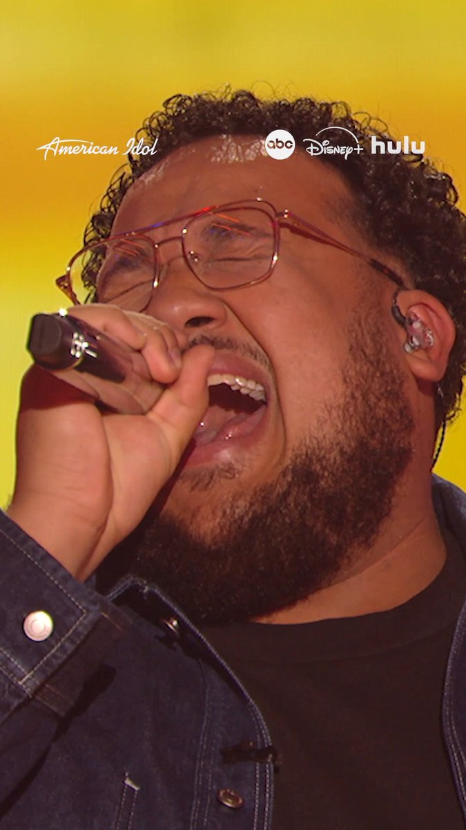 A close-up of a man with glasses and curly hair singing passionately into a microphone, with American Idol, ABC, Disney+, and Hulu logos displayed across the top.
