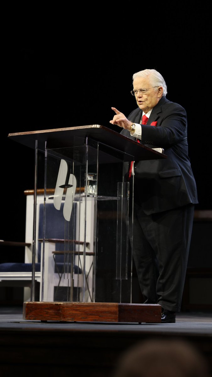 An elderly man with white hair, wearing a black suit, red tie, and glasses, stands behind a clear acrylic podium gesturing with his right hand as he delivers a sermon on a dark stage. The image aligns with the message about parents blessing their children, capturing a moment of passionate speaking that mirrors the call to "speak life" through simple acts of blessing.