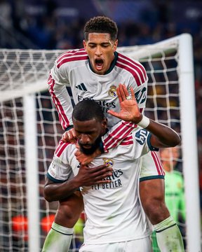 Two Real Madrid players in white away kits celebrate together, with one leaping onto the other's shoulders in an emotional embrace following a goal, capturing the intensity of a key match moment. The post notes Antonio Rüdiger's restrained celebration against his former club Roma, suggesting the jubilant teammate pictured is likely the goalscorer, while Rüdiger shows respect by holding back despite scoring the winner.