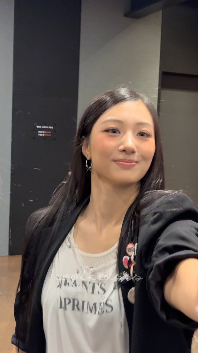 A selfie of a young woman with long dark hair smiling at the camera, wearing a black jacket over a white shirt with 'LOVE CHILD' and 'INFANTS' text visible, and several pins on her jacket lapel.
