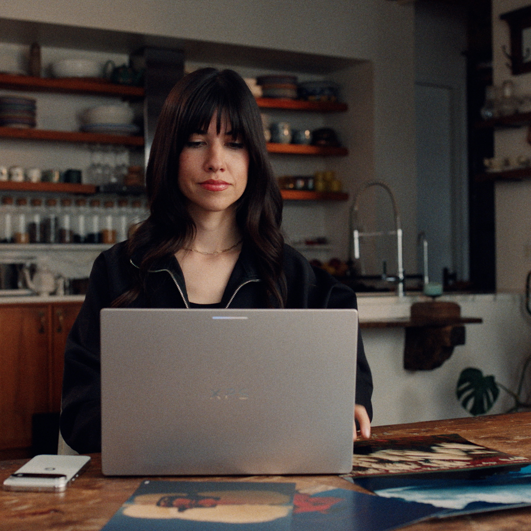 A woman works on a laptop at a wooden table in a warmly lit kitchen, with art prints and a phone beside her, embodying the seamless blend of creativity and AI-powered workflow described in the caption about Gemini and Googlebook.