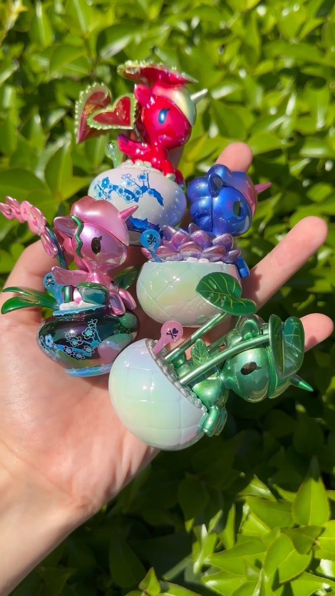 A hand holds a collection of small, colorful, iridescent Pop Mart toy characters in pink, blue, green, and purple hues against a leafy green background.