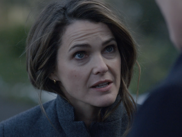 A woman with brown hair, blue eyes, and a serious expression stands outdoors in a gray coat, appearing to be in mid-dialogue with someone off-screen. The scene captures a tense, emotional moment that aligns with the dramatic finale of The Diplomat Season 3, teasing the highly anticipated Season 4 premiere.