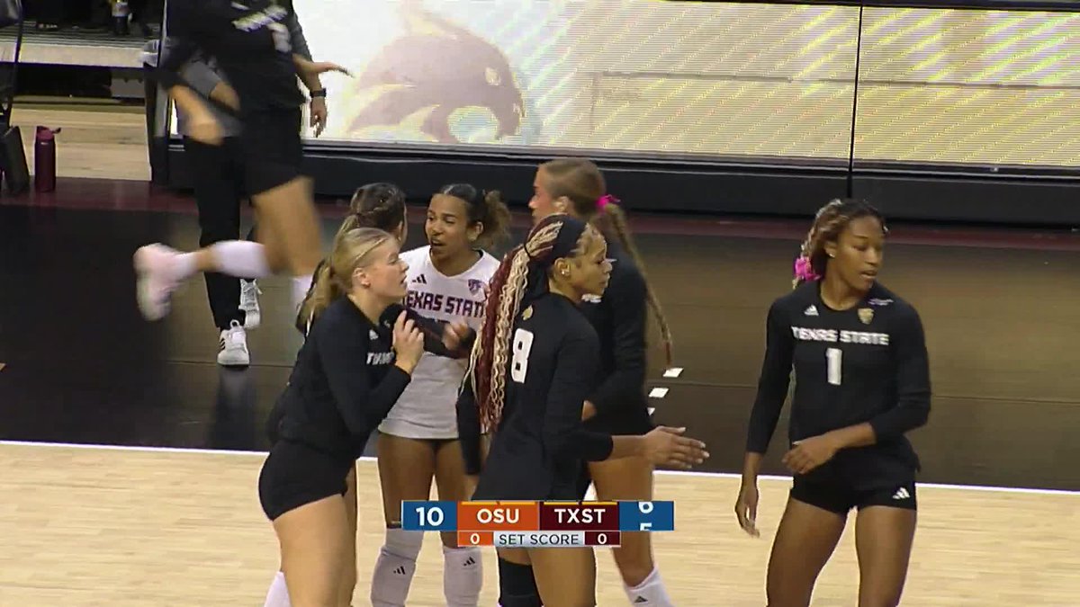 Texas State University volleyball players in black uniforms celebrate during a game, with the scoreboard showing OSU 10, TXST 0 in the first set.