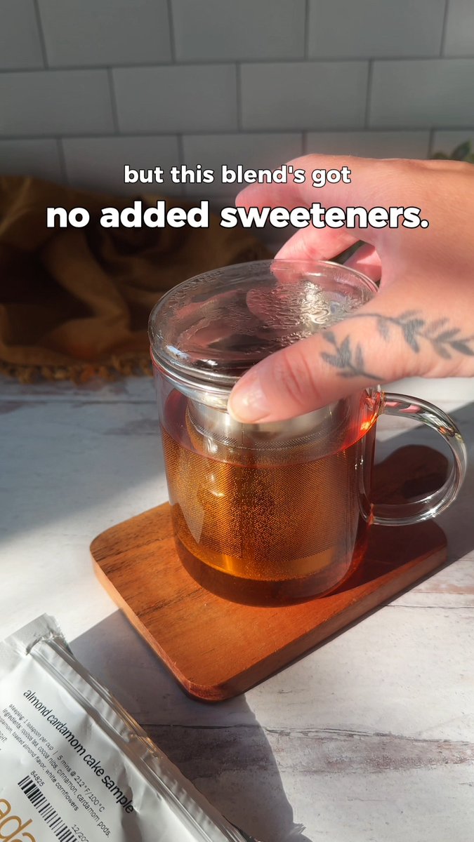 A hand with a leaf tattoo places a lid on a glass mug filled with tea, with text overlaid reading "but this blend's got no added sweeteners." while an almond cardamom cake sample packet rests nearby.