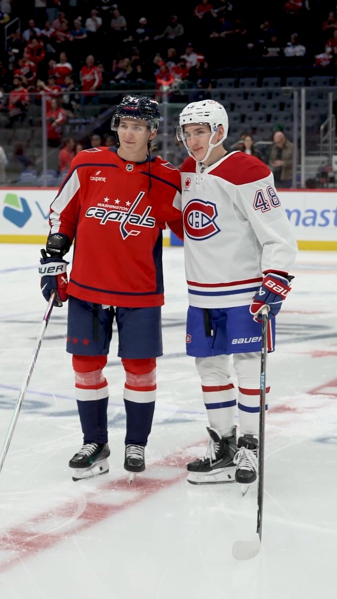 Two NHL players, both wearing helmets and full hockey gear, pose together on the ice — one in a red Washington Capitals jersey and the other in a white Montreal Canadiens jersey with the number 48. The image captures the "Hutson Bowl," the historic first NHL matchup between brothers, as referenced by the hashtag #GoHabsGo.